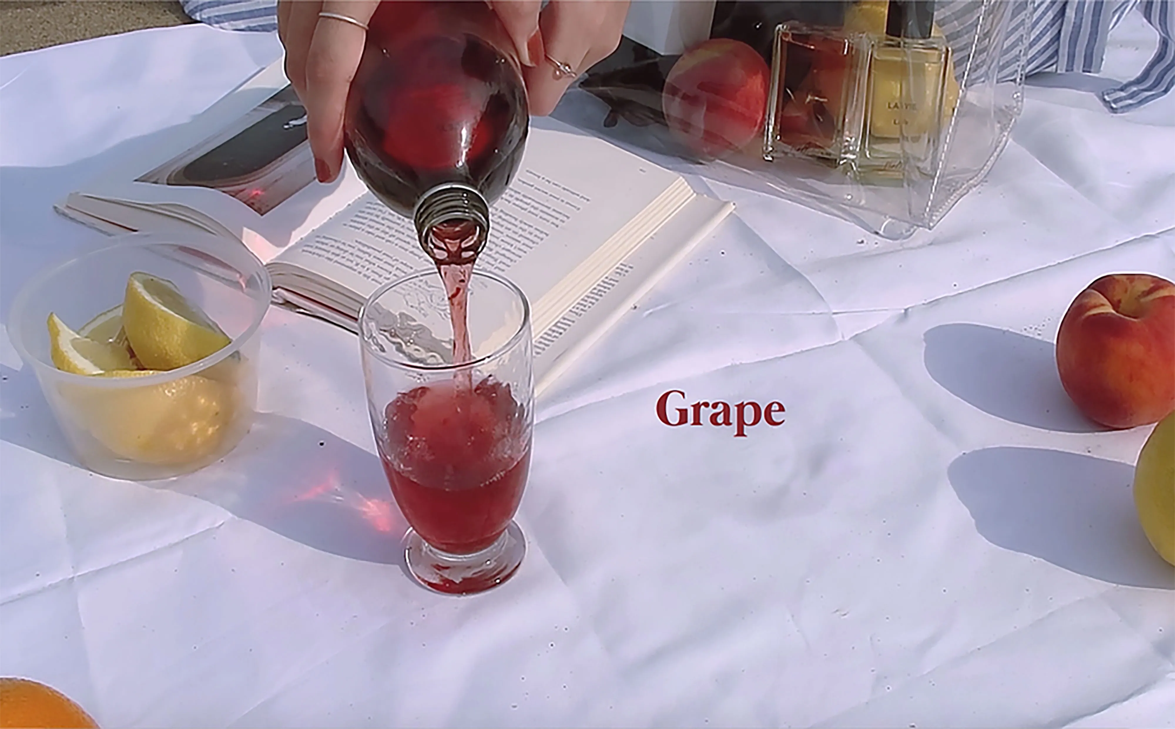 Video still: red juice poured into a glass on a picnic blanket, labelled Grape